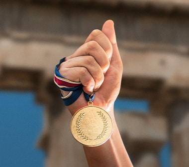 Hand holding gold medal in front of ancient Greek ruins.